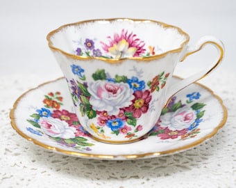 Può includere: Un set di tazza e piattino da tè bianchi con un design floreale. La tazza e il piattino hanno un bordo dorato e sono decorati con fiori rosa, rossi, blu e viola. La tazza ha un manico curvo.