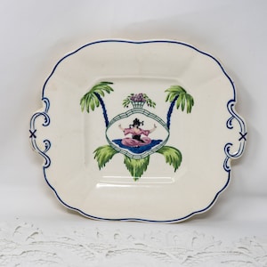 Könnte beinhalten: Ein weißer Keramik-Servierteller mit blauem und goldenem Rand. In der Mitte des Tellers befindet sich ein handgemaltes Design einer Frau in einem rosa Kleid, die zwischen zwei Palmen sitzt.