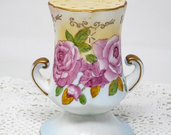 May include: A vintage porcelain salt shaker with a floral rose design. The shaker features a light yellow top with several holes, a white body with pink roses, green leaves, and gold accents. It has two small handles.