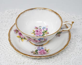 Puede incluir: Juego de taza y platillo blancos con un diseño floral de rosas rosas, moradas y azules. Los bordes son festoneados y ribeteados en dorado. La taza se encuentra sobre su platillo a juego.