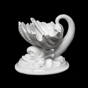 Puede incluir: Plato de cerámica blanca en forma de concha con una cuchara, sostenido por un diseño de delfín y olas. El plato tiene un cuello y una cola de cisne, y se asienta sobre una base redonda. La pieza es completamente blanca y tiene un acabado brillante.