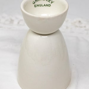 May include: White ceramic egg cup with a green 'Grindley England' inscription. The cup is designed to hold a single egg.