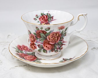 Puede incluir: Un juego de taza y platillo de té blanco con un patrón floral de rosas rosadas y hojas verdes. La taza y el platillo tienen un borde dorado.