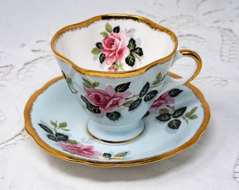May include: A delicate teacup and saucer set with a light blue background and a floral pattern of pink roses and green leaves. The edges of the cup and saucer are trimmed with gold.