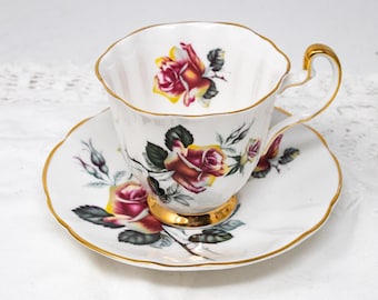 May include: A white teacup and saucer with a gold rim. The cup and saucer are decorated with a floral pattern of red, yellow, and green roses.