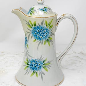 Peut inclure: Une théière en porcelaine blanche avec une bordure dorée et un motif floral bleu. La théière a un couvercle et une poignée.