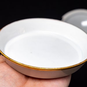May include: A white porcelain dish with a gold rim and two gold handles. The dish is empty and has a slight curve to the bottom.