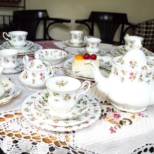Op de afbeelding: Een wit theeservies met bloemenmotieven, inclusief een theepot, roomkan, suikerpot en kopjes en schoteltjes. De set is gerangschikt op een wit tafelkleed met een gehaakt kanten overtrek. Een plak cake met rode kersen staat op een bord in het midden van de tafel.