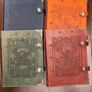 May include: Four faux leather bound journals, each featuring a different Hogwarts house crest. The crests are for Gryffindor, Slytherin, Hufflepuff, and Ravenclaw. Each journal has a gold clasp and decorative stitching.