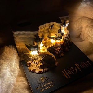May include: A 3D pop-up book shaped like Hogwarts castle from the Harry Potter series. The book is lit from within, illuminating the castle's details. The book's cover is black with the words "Wizarding World" and "Harry Potter" in gold lettering.
