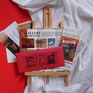 May include: A red envelope with a stamp that says "SEVI" on a wooden easel. The easel is surrounded by brochures with images of artwork and the text "GALLERY HIGHLIGHTS" and "SCREAMING VENICE".