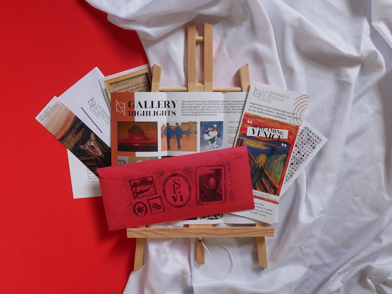 May include: A red envelope with a stamp that says "SEVI" on a wooden easel. The easel is surrounded by brochures with images of artwork and the text "GALLERY HIGHLIGHTS" and "SCREAMING VENICE".