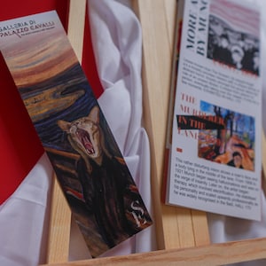 May include: A bookmark featuring a cat with an open mouth, a parody of Edvard Munch's "The Scream". The bookmark is on a wooden easel with a white cloth background. The text on the bookmark reads "Galleria di Palazzo Cavalli".