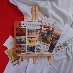 May include: A white easel displaying a gallery highlights brochure featuring a variety of artwork, including a painting of a screaming figure, a painting of a man in a field, and a painting of a sunset. The brochure also includes the text "Gallery Highlights" and "Edward Munch".
