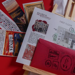 May include: A red envelope with a black stamp and the text "LE VI" on it. The envelope is on top of a wooden easel with a map of the first floor of the Galleria di Palazzo Cavalli, a museum in Venice, Italy. The map shows the location of various rooms and exhibits. The map is printed on white paper with black text and symbols. The map is surrounded by other items, including a bookmark with a painting of a screaming figure, a bookmark with a painting of a cat, and a bookmark with a polka dot pattern.