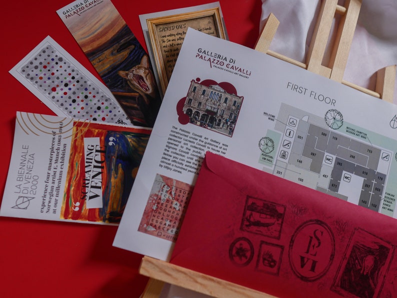 May include: A red envelope with a black stamp and the text "LE VI" on it. The envelope is on top of a wooden easel with a map of the first floor of the Galleria di Palazzo Cavalli, a museum in Venice, Italy. The map shows the location of various rooms and exhibits. The map is printed on white paper with black text and symbols. The map is surrounded by other items, including a bookmark with a painting of a screaming figure, a bookmark with a painting of a cat, and a bookmark with a polka dot pattern.