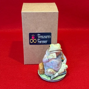 May include: A ceramic frog figurine in a light purple coat, holding a card, on a green base. A brown box with a "Treasures Forever" sticker is behind it. The background is red. The figurine is a decorative item.