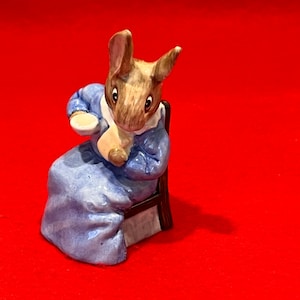 May include: A small ceramic figurine of a rabbit wearing a blue dress and holding a cup. The rabbit is sitting on a wooden chair.