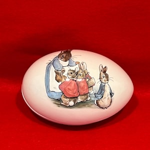 May include: A white ceramic egg-shaped trinket box with a painted image of Peter Rabbit and his siblings. The box features a blue background with a red and white design.