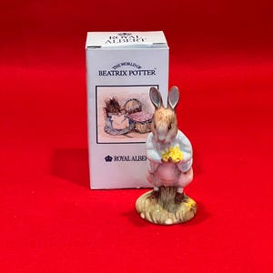 May include: A Royal Albert ceramic figurine of a rabbit from "The World of Beatrix Potter". The rabbit wears a blue jacket and pink skirt, holding yellow flowers. The figurine stands on a grassy base, with a box featuring illustrations.
