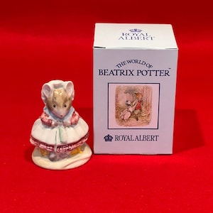 May include: A ceramic mouse figurine in a dress and bonnet, with a box beside it. The box is white and features the text "THE WORLD of BEATRIX POTTER" and the Royal Albert logo. The figurine sits on a red surface.