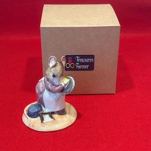 May include: A ceramic mouse figurine wearing a white apron and holding a small frying pan with a yellow piece of food. The mouse stands on a circular base next to a small dustpan. Behind the figurine is a square cardboard box with a sticker reading Treasures Forever.