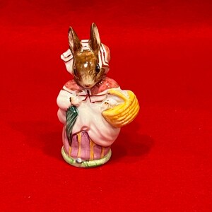 May include: A ceramic rabbit figurine wearing a white and pink outfit, carrying a yellow basket and a green umbrella. The rabbit has brown ears and a white bonnet. The figurine has a striped skirt and stands on a green base. The figurine is approximately 10 cm tall.