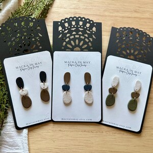 May include: Three pairs of handmade clay earrings in various colors, displayed on white cards with black decorative cutouts. Each earring set features a unique combination of shapes and hues, including black, white, brown, and olive green. The text "MACRA IN MAY" is visible.