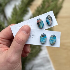 May include: Oval-shaped stud earrings with a marbled design in shades of blue, pink, gold, and black. The earrings are displayed on white cards. The earrings are held by a person's hand.