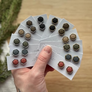May include: A hand holds several cards displaying hexagon-shaped stud earrings in various colors. The earrings have a textured appearance and are in shades of black, green, tan, and red. Some earrings have silver or gold-colored borders.