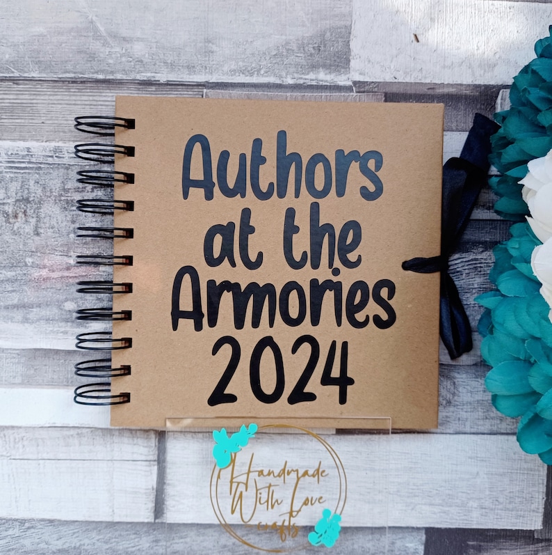 May include: A brown scrapbook with a black spiral binding. The cover has the text "Authors at the Armories 2024" in black lettering. A black ribbon is tied around the book.