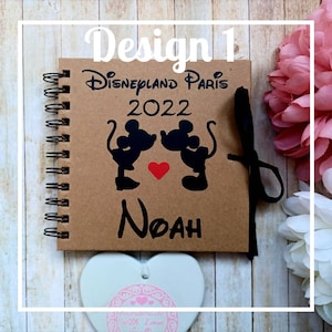 May include: A brown notebook with a spiral binding. The cover features a black silhouette of Mickey Mouse and Minnie Mouse kissing with a red heart. The text "Disneyland Paris 2022" and the name "Noah" are printed on the cover.