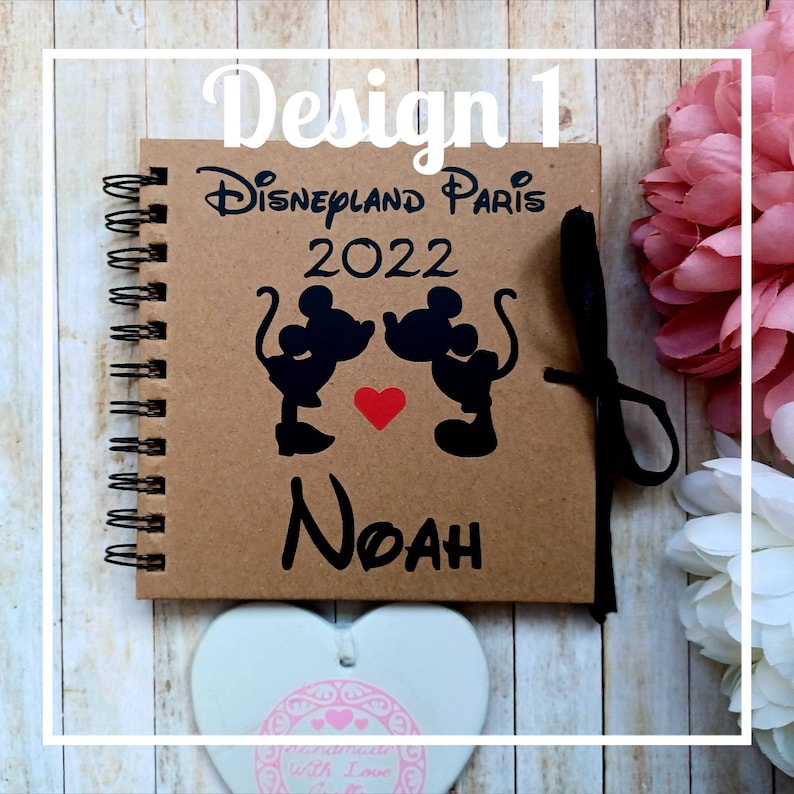 May include: A brown notebook with a spiral binding. The cover features a black silhouette of Mickey Mouse and Minnie Mouse kissing with a red heart. The text "Disneyland Paris 2022" and the name "Noah" are printed on the cover.