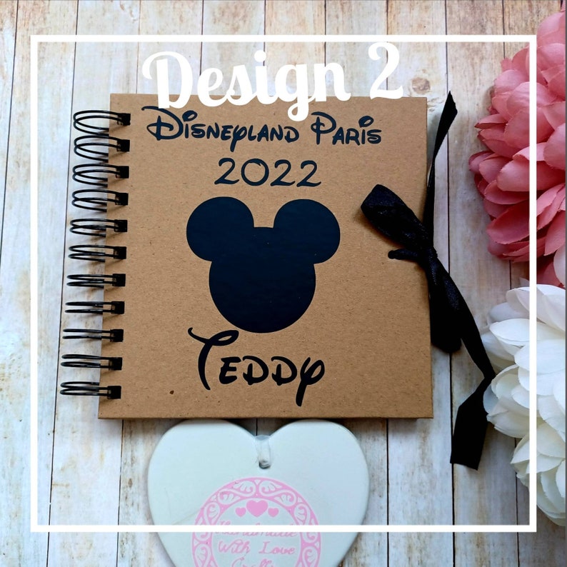 May include: A brown scrapbook with a black Mickey Mouse head and the text "Disneyland Paris 2022 Teddy" on the cover. The scrapbook has a black ribbon tied around it.