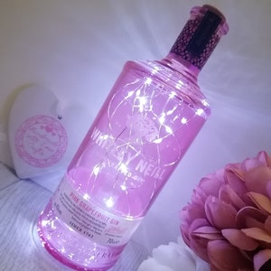 May include: A pink glass bottle with white fairy lights wrapped around it. The bottle is filled with a pink liquid and has a label that reads "Whirlley Neill Pink Grapefruit Gin" and "Since 1762".