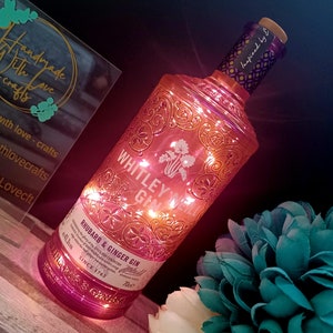 May include: A pink glass bottle with a gold design and a label that reads "Whitley Neill Gin Rhubarb & Ginger Gin" with a small illustration of a rhubarb plant. The bottle is lit with fairy lights and has a cork stopper with a purple and gold label that reads "Inspired by E".