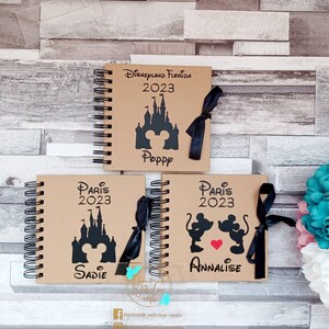 May include: Three brown notebooks with black spiral bindings. Each notebook has a black and white illustration of a Disney castle and Mickey Mouse. The top notebook says "Disneyland Florida 2023" and "Poppy". The middle notebook says "Paris 2023" and "Sadie". The bottom notebook says "Paris 2023" and "Annalise".
