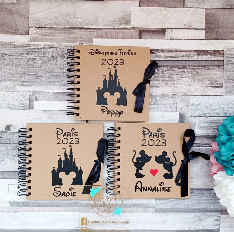 May include: Three brown notebooks with black spiral bindings. Each notebook has a black and white illustration of a Disney castle and Mickey Mouse. The top notebook says "Disneyland Florida 2023" and "Poppy". The middle notebook says "Paris 2023" and "Sadie". The bottom notebook says "Paris 2023" and "Annalise".