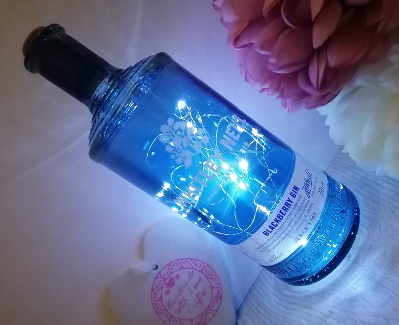 May include: A blue glass bottle with a white label that says "Blackberry Gin" and "Whitley Neill" with white fairy lights wrapped around it. The bottle is sitting on a white surface.