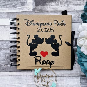 May include: A brown scrapbook with a black spiral binding. The cover features a Mickey Mouse and Minnie Mouse silhouette with a red heart and the text "Disneyland Paris 2025" and "Rory".