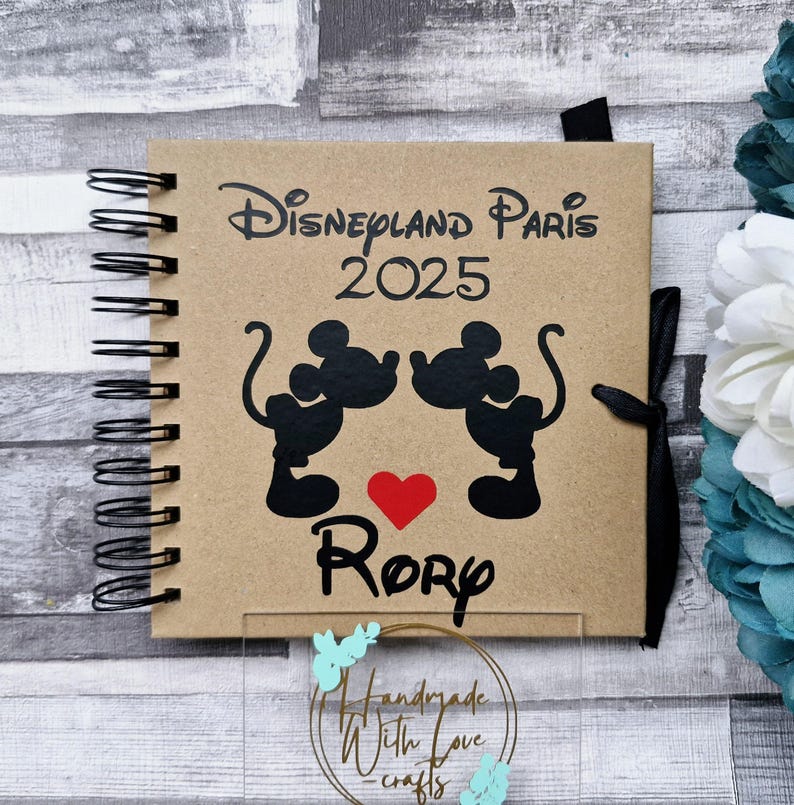 May include: A brown scrapbook with a black spiral binding. The cover features a Mickey Mouse and Minnie Mouse silhouette with a red heart and the text "Disneyland Paris 2025" and "Rory".