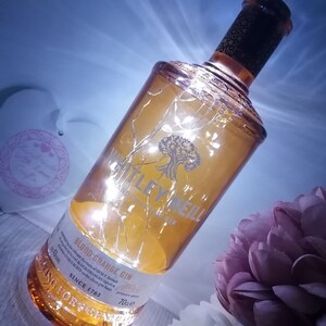 May include: A clear glass bottle of Whitley Neill Blood Orange Gin with a gold label and a string of white fairy lights wrapped around it. The bottle is sitting on a white surface with a pink flower in the background.