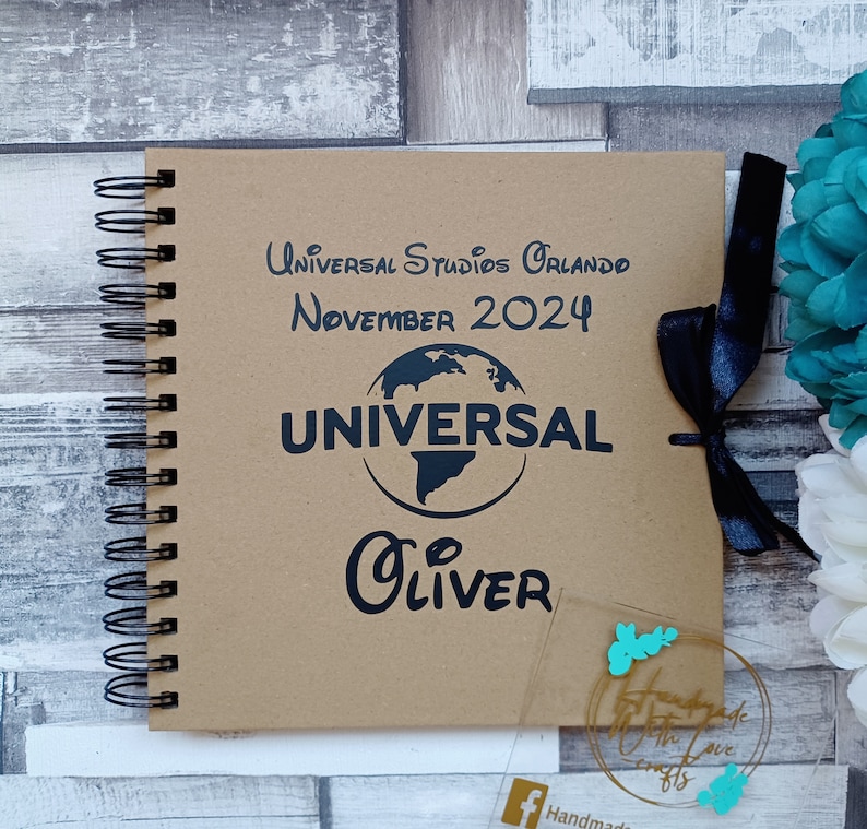 May include: A brown scrapbook with a black spiral binding. The cover has the text "Universal Studios Orlando November 2024" and a black globe with the word "Universal" in white text. The name "Oliver" is written in black cursive below the globe. A black ribbon is tied around the book.