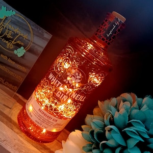 May include: A clear glass bottle with a textured surface and a label that reads "Whitley Neill Gin" and "Raspberry Gin". The bottle is illuminated with warm white fairy lights and has a cork stopper.