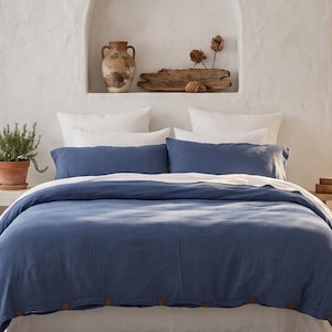May include: A bed featuring a deep blue duvet cover with visible brown button details, layered over white bedding. The bed is positioned in a room with white walls, a central arched wall niche, sheer curtains, and a potted olive tree on the right.