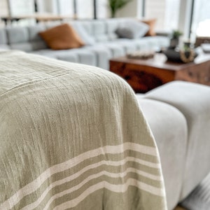 May include: A sage green throw blanket with three wide, horizontal, off-white stripes. The blanket has a textured weave and a fringed edge. The blanket is draped over a gray sofa, suggesting a cozy home decor theme.