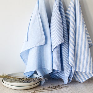 May include: Three blue kitchen towels hanging on hooks. Two towels have a white stripe pattern, and one towel is a solid light blue color.