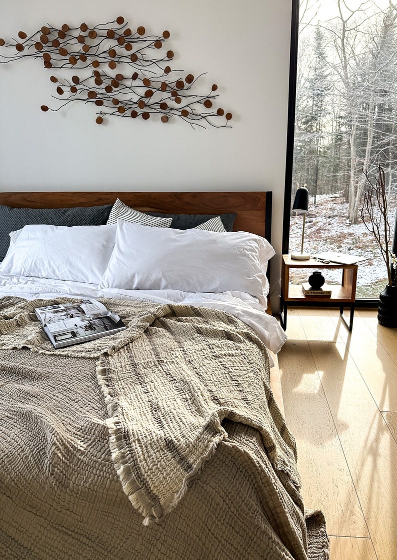 May include: A bedroom with a wooden bed frame, white bedding, and a beige throw blanket. The bed is next to a window with a view of a snowy forest. A small wooden side table with a lamp and a book is next to the bed.