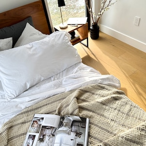May include: A white bed with a white duvet cover and a striped throw blanket. A magazine is open on the blanket. A small wooden side table with a black lamp and a laptop is next to the bed.