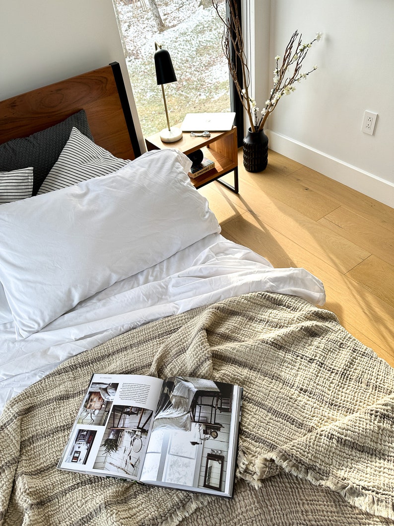 May include: A white bed with a white duvet cover and a striped throw blanket. A magazine is open on the blanket. A small wooden side table with a black lamp and a laptop is next to the bed.